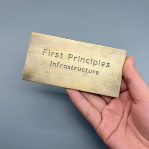 May include: A rectangular, brass-colored plaque with the words "First Principles" and "Infrastructure" engraved in a dark font. The plaque is held in a hand against a gray background.
