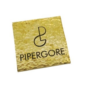 May include: A square, textured, gold-coloured metal plate with the name "PIPERGORE" in black, sans-serif font. The plate also features a black logo. The surface has a hammered appearance, and the background is white.