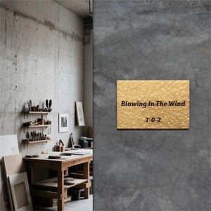 May include: A gold-coloured rectangular plaque with the text "Blowing In The Wind" and "1-0-2" on a grey concrete wall. The plaque has a textured surface. The background shows a workshop with tools and a wooden table.