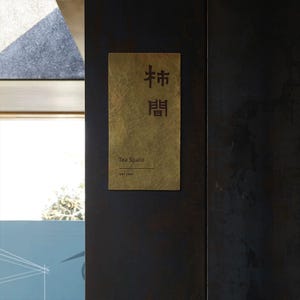 Op de afbeelding: Een rechthoekig messing bord met de woorden "Tea Space" en Chinese karakters in zwart. Het bord is bevestigd aan een donkerbruine muur. De woorden "Shi Jian" staan onder de woorden "Tea Space".