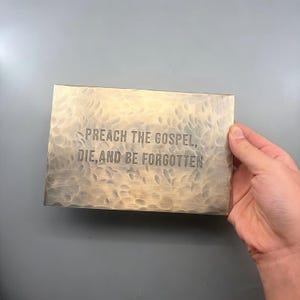 以下が含まれることがあります： 「PREACH THE GOSPEL, DIE, AND BE FORGOTTEN」の文字が刻まれた、ハンマー加工が施された長方形の金属板。 金色の色合いで、灰色の背景に手で持たれています。