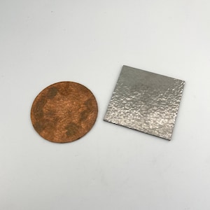 May include: A copper-colored, round coaster with a textured surface sits next to a square, silver-colored coaster with a hammered texture. Both coasters are displayed on a white background.