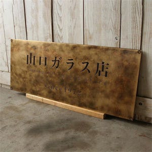 May include: A rectangular brass sign with the text "山口ガラス店 Yamaguchi glass store since 1972" in black lettering. The sign is mounted on a wooden base and has a weathered appearance.