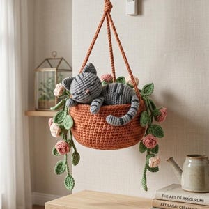 May include: A crocheted hanging planter with a gray striped cat nestled inside. The orange basket is adorned with pink and green crocheted flowers and leaves. The planter hangs from a brown rope attached to a white bracket on a beige wall.