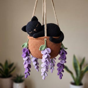 Könnte beinhalten: Ein handgefertigter hängender Häkelkorb mit einer schlafenden schwarzen Katze darin. Der braune Korb ist mit lila und grünen Häkelblumen verziert, die an beigen Schnüren aufgehängt sind. Die Katze hat geschlossene Augen.
