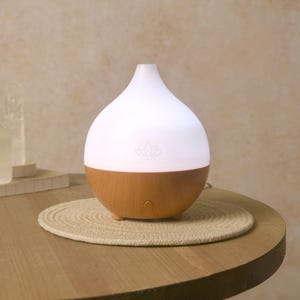 May include: A white and brown essential oil diffuser with a lotus flower logo. The top half is white and illuminated, while the bottom half is wood-toned. It sits on a round, woven mat on a wooden table.