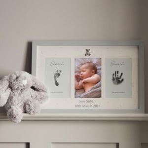 May include: A baby photo frame in grey, featuring a central photo of a sleeping baby, with prints of a foot and hand on either side. The frame includes the baby's name and birthdate. A grey plush bunny is on the left.
