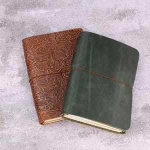 May include: Two leather journals. One is brown with a floral embossed design and a brown elastic closure. The other is dark green with a brown elastic closure. The journals are open, revealing the pages.