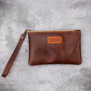 May include: A brown leather wristlet with a gold zip and a matching wrist strap. The wristlet features the name "Simon" embossed on a rectangular patch. The leather has a distressed appearance.