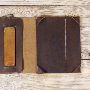 May include: A brown leather notebook cover with a flap closure and a built-in pen holder. The cover has a distressed look and is stitched with brown thread. The notebook cover is open to reveal a tan leather interior.