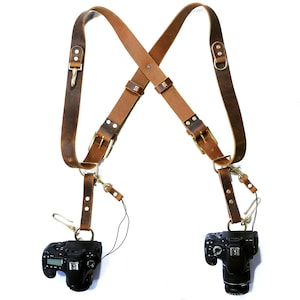 May include: A brown leather camera strap with two adjustable straps and two metal D-rings. The strap is designed to hold two cameras, one on each side. The cameras are black and have a lens on the front.