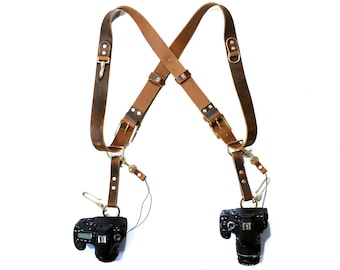 May include: A brown leather camera strap with two adjustable straps and two metal D-rings. The strap is designed to hold two cameras, one on each side. The cameras are black and have a lens on the front.