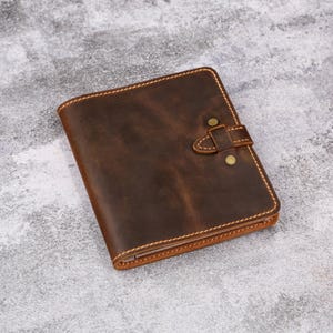 May include: A brown leather wallet with a buckle closure and brass-colored rivets. The wallet has a stitched border and a distressed finish, suggesting a vintage or rustic style. The wallet is approximately 4 inches wide and 5 inches tall.