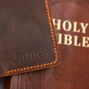 May include: Close-up of a brown leather item with the name "James" embossed on it. The item has a stitched border with light brown thread. A portion of a brown leather Bible with the words "Holy Bible" is visible in the background.