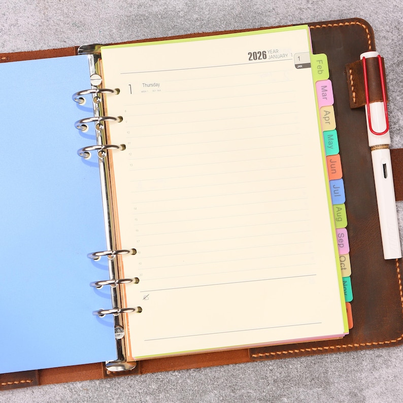 May include: A brown leather binder with a six-ring metal mechanism holding cream-colored 2026 calendar pages. The planner features colorful monthly index tabs and a white fountain pen with a red clip stored in a leather loop on the right side.
