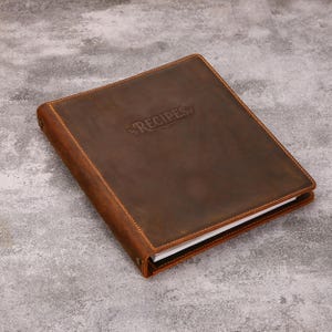 Puede incluir: Un archivador de recetas de cuero marrón oscuro con un acabado mate liso y costuras visibles a lo largo de los bordes. La palabra RECIPES está grabada en el centro de la portada y el archivador contiene hojas de papel blanco visibles en el lateral.