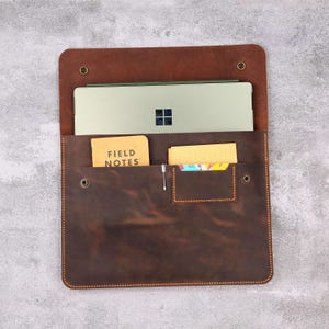 May include: A brown leather laptop sleeve with a top flap secured by snaps. The sleeve has a pocket for a notepad with "FIELD NOTES" printed on it, a pen, and a card. A silver laptop is inside the sleeve.