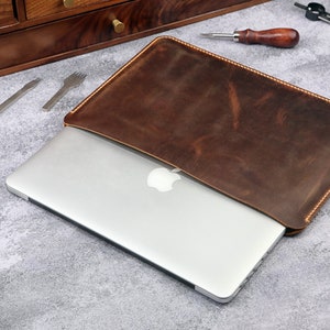 May include: A brown leather laptop sleeve with a stitched edge. The sleeve is open and a silver laptop is inside. The laptop has an Apple logo on the back.