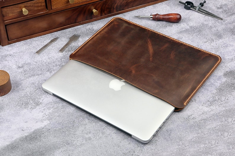 May include: A brown leather laptop sleeve with a stitched edge. The sleeve is open and a silver laptop is inside. The laptop has an Apple logo on the back.