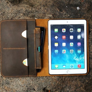 May include: A brown leather folio case with a flap closure and a built-in pocket for a tablet or phone. The case has a brown leather interior and a brown leather strap. The case is open and a tablet is visible inside. The tablet has a white screen with a blue background and a variety of icons.