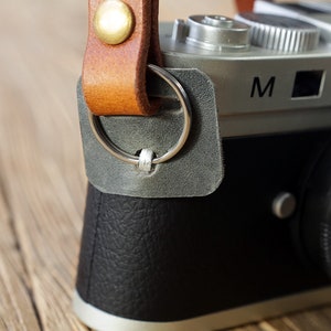 May include: A silver camera with a black leather strap and a black lens. The camera has a silver ring around the lens and the letter "M" on the top.
