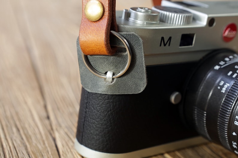 May include: A silver camera with a black leather strap and a black lens. The camera has a silver ring around the lens and the letter "M" on the top.