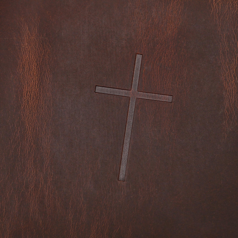 May include: Close-up of a brown leather surface with a debossed cross. The leather has a textured appearance with variations in color, ranging from light to dark brown. The cross is centered and has a simple, clean design.