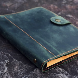 May include: A dark green leather bound journal with a flap closure and a brass button. The journal has orange stitching and is lying on a textured gray surface.