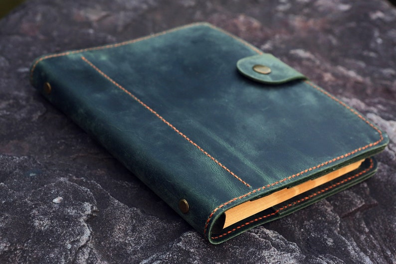 May include: A dark green leather bound journal with a flap closure and a brass button. The journal has orange stitching and is lying on a textured gray surface.