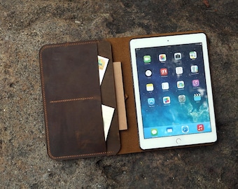 May include: A brown leather iPad case with a white iPad inside. The case has a pocket for cards and a pen holder. The iPad screen displays app icons. The case is open and resting on a textured, gray surface.