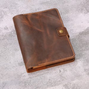 May include: A dark brown leather notebook cover featuring a smooth, distressed texture with visible grain patterns. The cover is secured by a matching leather strap with a single brass-colored metal snap button closure and thick, light-colored stitching.