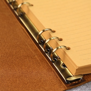 May include: A brown leather binder featuring a metallic bronze-toned six-ring mechanism. The interior contains light tan lined paper pages held securely by the circular metal rings, which are attached to the textured brown cover material.