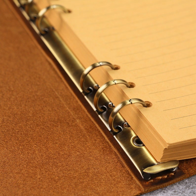 May include: A brown leather binder featuring a metallic bronze-toned six-ring mechanism. The interior contains light tan lined paper pages held securely by the circular metal rings, which are attached to the textured brown cover material.