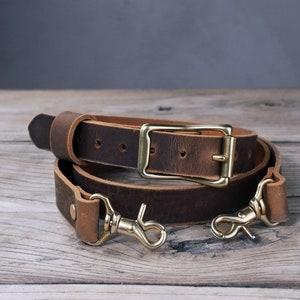 May include: A brown leather camera strap with a brass buckle and two brass hooks.