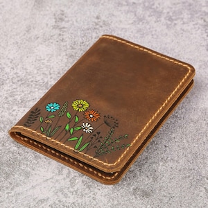 Puede incluir: Una cartera de cuero marrón con un diseño pintado a mano de flores silvestres coloridas, incluyendo pétalos amarillos, naranjas, blancos y azules con tallos y hojas verdes, rematada con costuras gruesas de color tostado en los bordes de la cubierta.