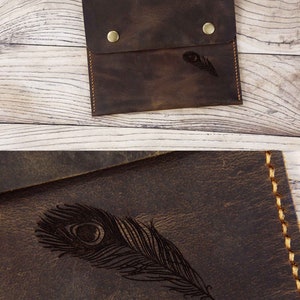 Puede incluir: Una bolsa de cuero marr&oacute;n con cierre de solapa y dos broches de lat&oacute;n. La bolsa tiene un dise&ntilde;o de pluma de pavo real grabado en la parte delantera.