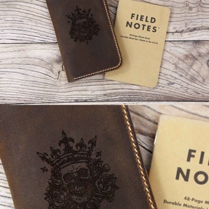 May include: A dark brown leather notebook cover featuring a laser-engraved skull wearing a crown surrounded by roses, with visible tan stitching along the edges, paired with a tan Field Notes 48-page memo book with black text.