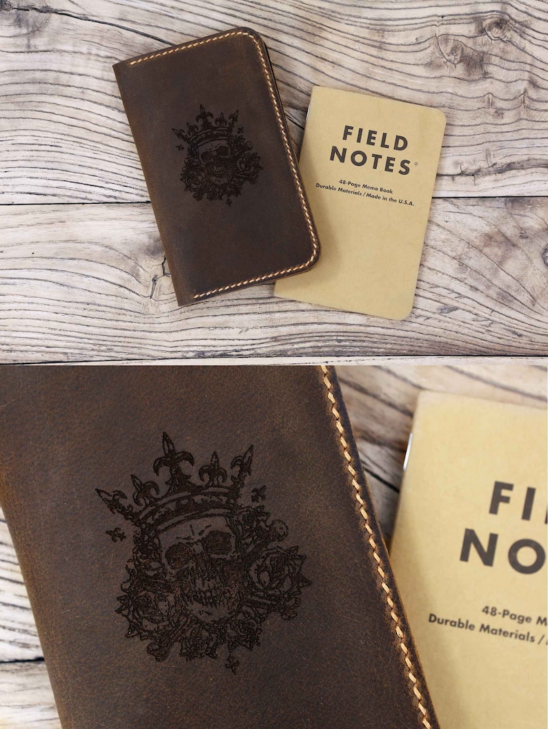 May include: A dark brown leather notebook cover featuring a laser-engraved skull wearing a crown surrounded by roses, with visible tan stitching along the edges, paired with a tan Field Notes 48-page memo book with black text.