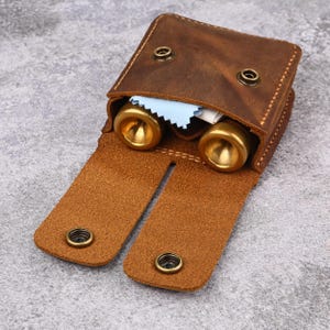 Puede incluir: Un estuche de cuero marrón con dos objetos de latón en su interior. El estuche está abierto, revelando los objetos y un paño de limpieza azul. El estuche tiene dos cierres de botón y es de cuero texturizado.