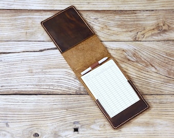 May include: A brown leather notepad holder with a pen and lined paper inside. The holder is open and the paper is visible. The holder is on a wooden surface.