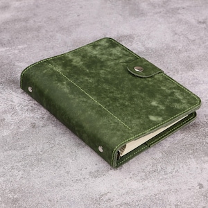 May include: A rectangular notebook with a textured, forest green cover featuring visible stitching along the edges. The notebook includes a flap closure secured by a single silver-toned metal snap button and two silver-toned rivets on the spine.