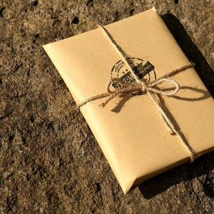Puede incluir: Un paquete de papel marr&oacute;n atado con cuerda y sellado con un sello redondo de cera negra. El sello tiene el texto "Original" y "Handmade" dentro de un c&iacute;rculo.