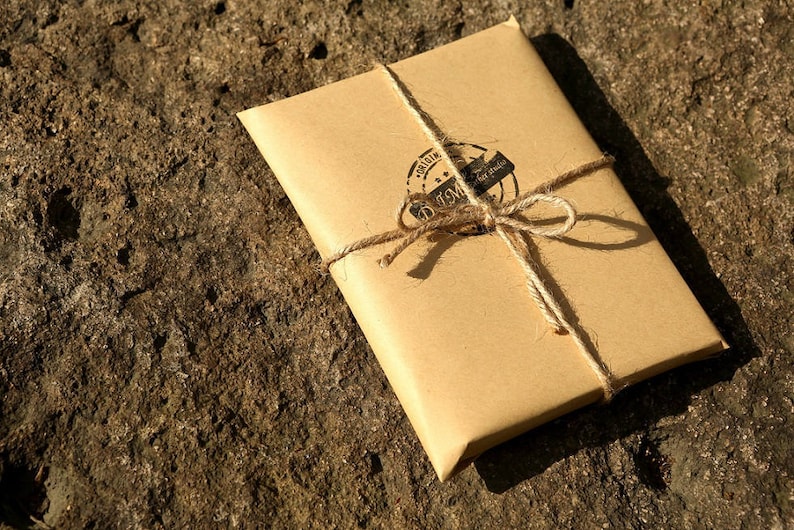 Puede incluir: Un paquete de papel marr&oacute;n atado con cuerda y sellado con un sello redondo de cera negra. El sello tiene el texto "Original" y "Handmade" dentro de un c&iacute;rculo.