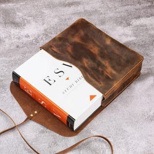 May include: A brown leather Bible cover with a distressed finish, holding a white book with the title "ESV STUDY BIBLE" in black and orange lettering. The cover has a flap closure and a long strap.