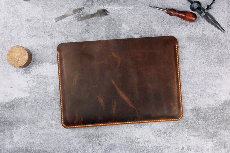 May include: A brown leather laptop sleeve with a stitched edge. The sleeve is made of distressed leather and has a rustic look.