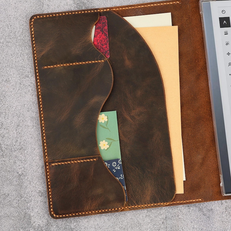Op de afbeelding: Een donkerbruine lederen hoes voor een e-reader met een gebogen binnenzakontwerp en zichtbare oranje stiksels. De zak bevat een rood patroonkaartje, een groene kaart met gele bloemen, een blauwe bloemenkaart en een beige papieren document.