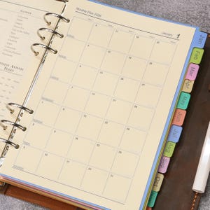 May include: A ring-bound planner open to a January 2026 monthly calendar page with cream-colored paper. The planner features colorful monthly index tabs along the right edge and a list of flowers and Chinese animal years on the left page.