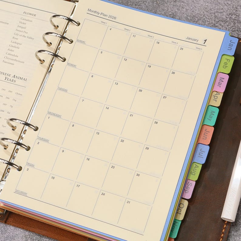 May include: A ring-bound planner open to a January 2026 monthly calendar page with cream-colored paper. The planner features colorful monthly index tabs along the right edge and a list of flowers and Chinese animal years on the left page.