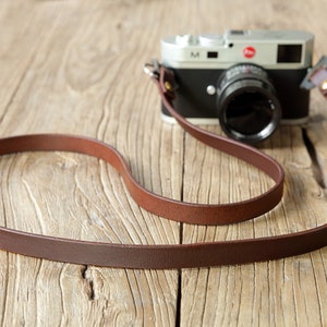 May include: A brown leather camera strap with a silver camera attached. The strap is lying on a wooden surface.