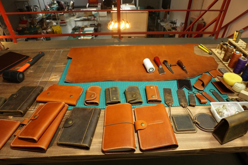 May include: A workbench with various leather goods, including wallets, keychains, and notebooks. There is a large piece of brown leather on the workbench, along with tools for leatherworking.